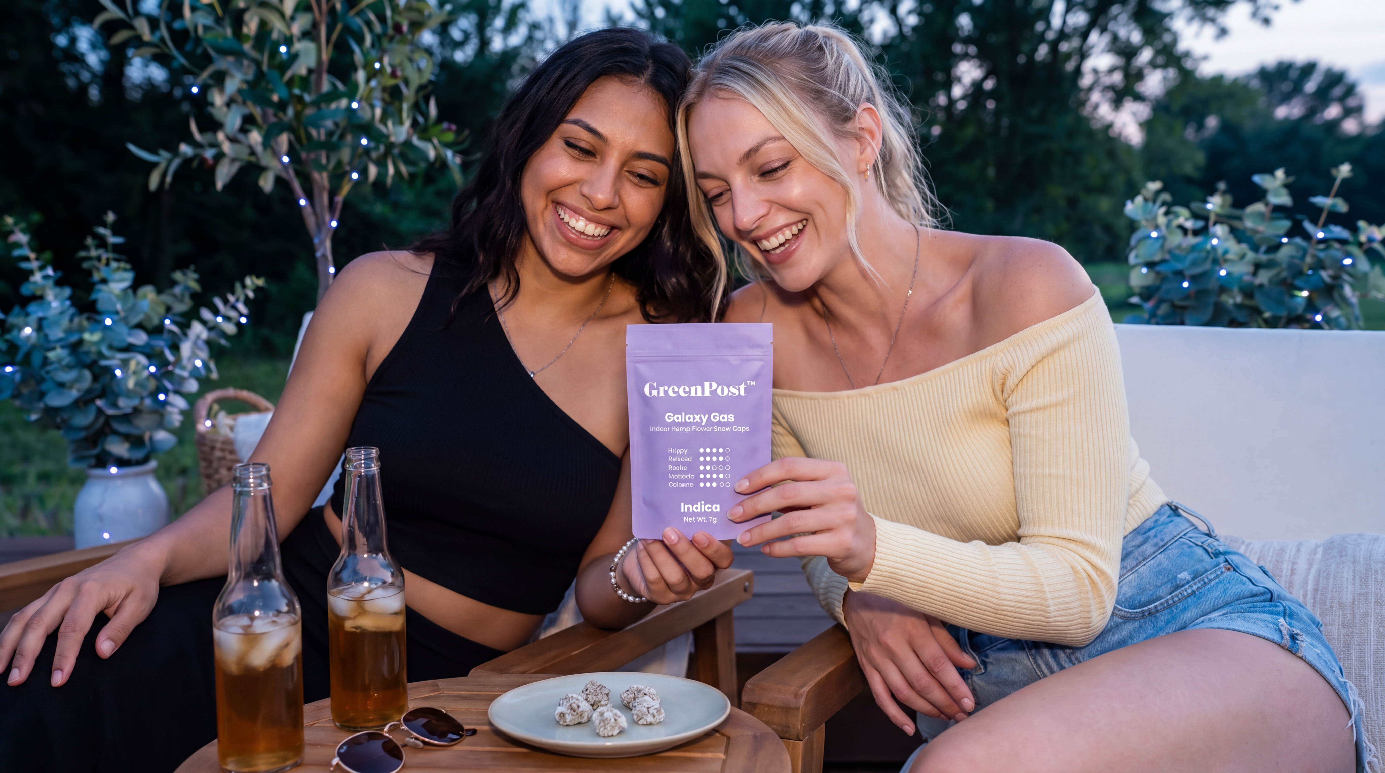 Two adult women sitting outdoors at dusk holding a GreenPost Galaxy Gas Snow Caps pouch with frosted THCA flower on a plate.