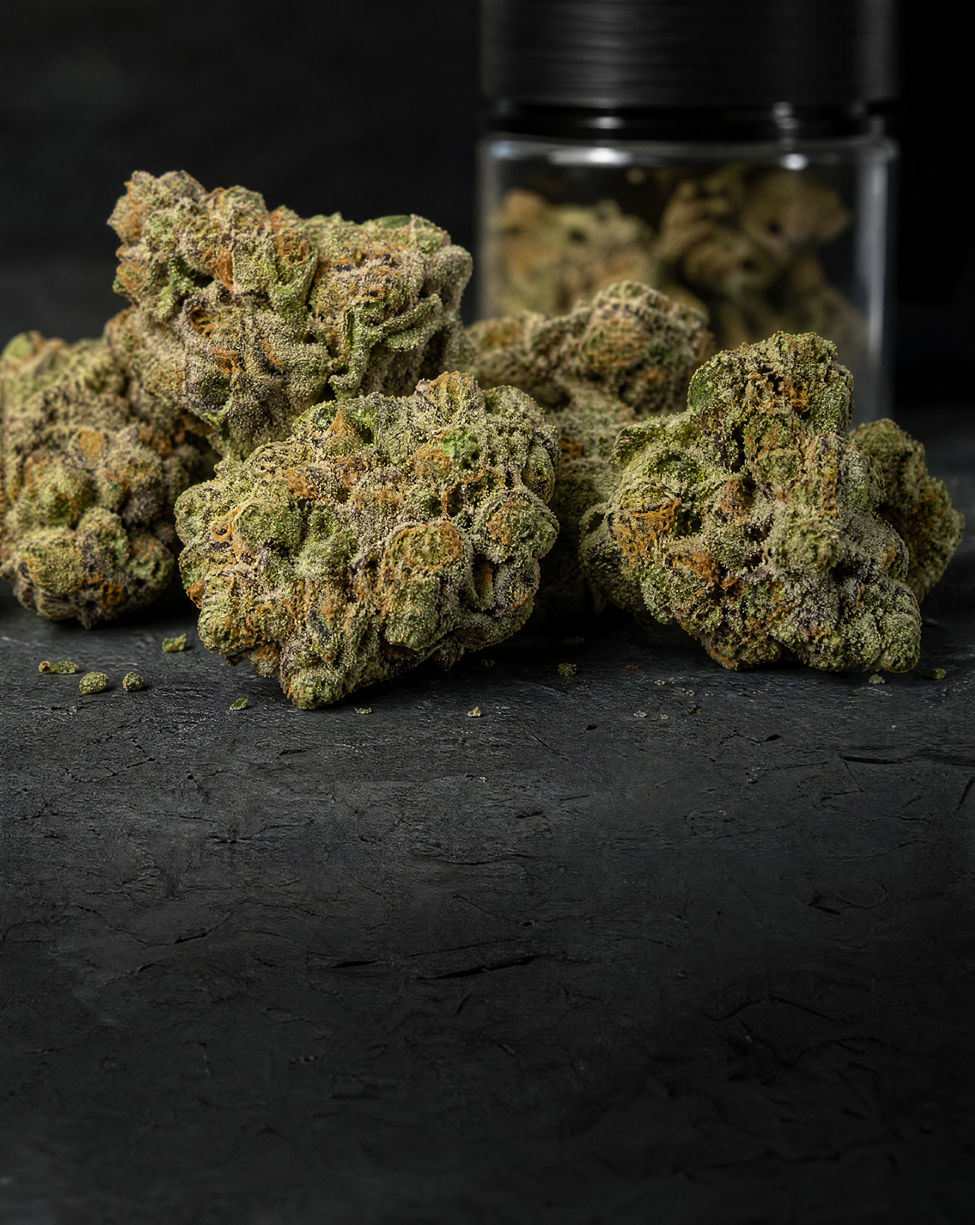 Close-up of dense, frosty indoor THCA flower buds on a dark stone surface with a glass jar blurred in the background.