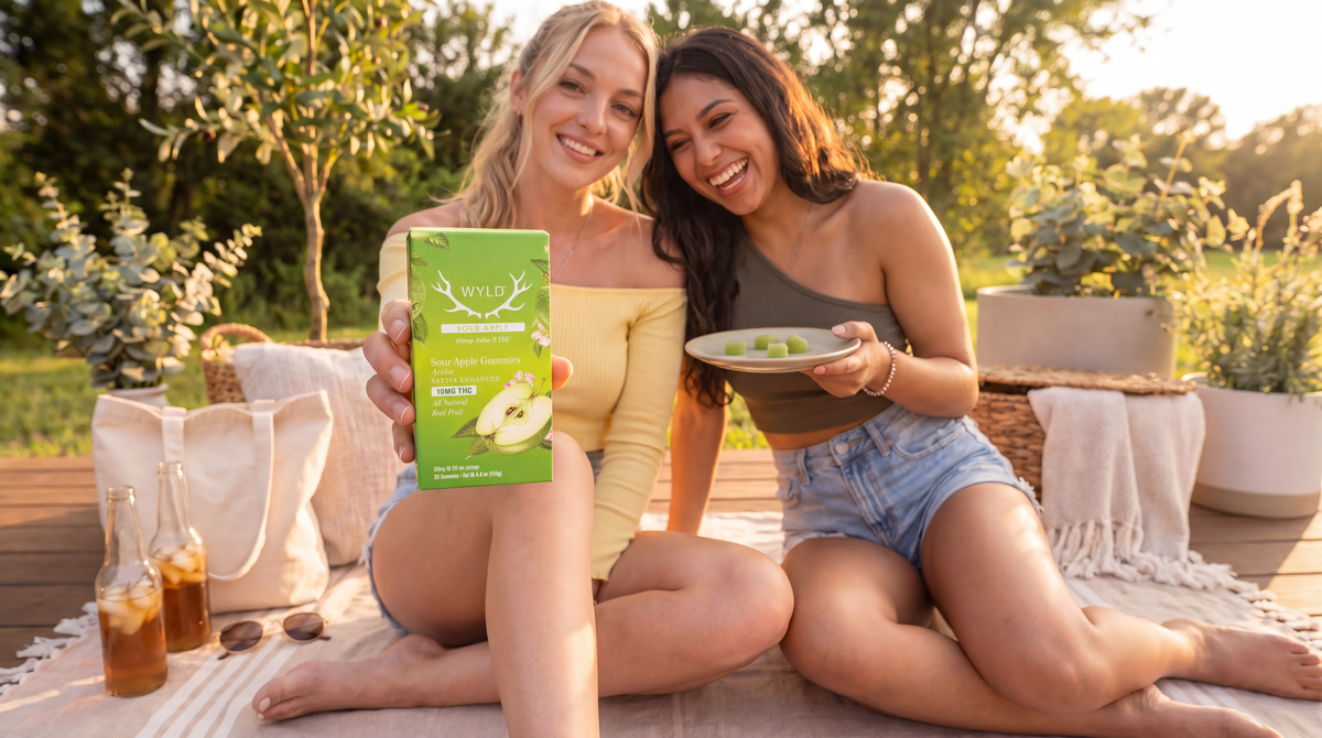 Two adult women sitting on an outdoor deck holding a Wyld Sour Apple Delta 9 THC Gummies box and green gummies on a plate.
